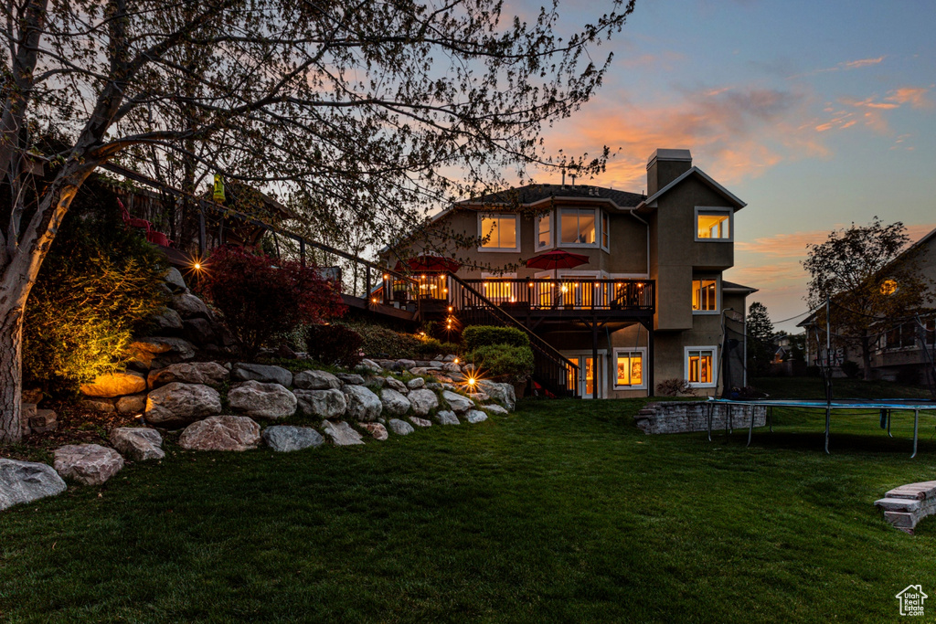 Back of property at dusk with a wooden deck, a trampoline, a chimney, and a yard
