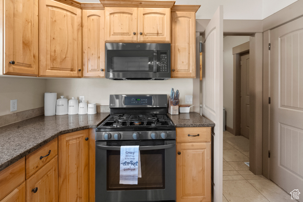 Kitchen featuring black microwave, stainless steel gas stove, dark stone countertops, light brown cabinets, and light tile patterned flooring