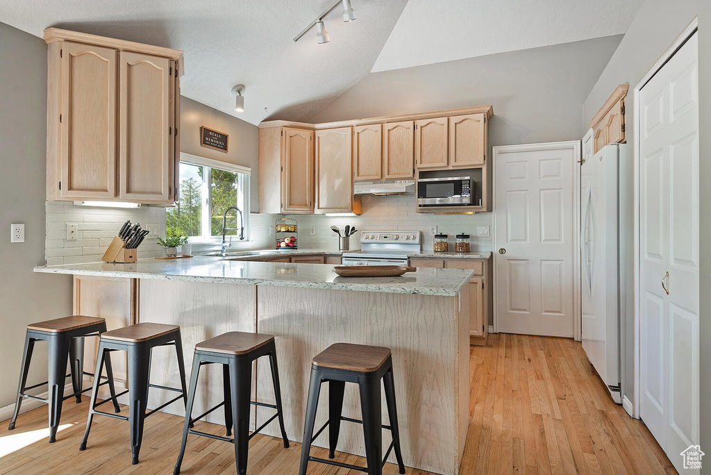 Kitchen featuring a peninsula, light brown cabinetry, white appliances, a sink, and vaulted ceiling