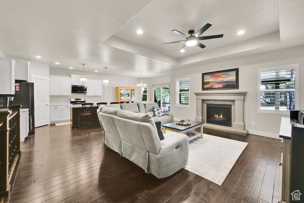 Living room with dark wood finished floors, a raised ceiling, ceiling fan with notable chandelier, a warm lit fireplace, and recessed lighting