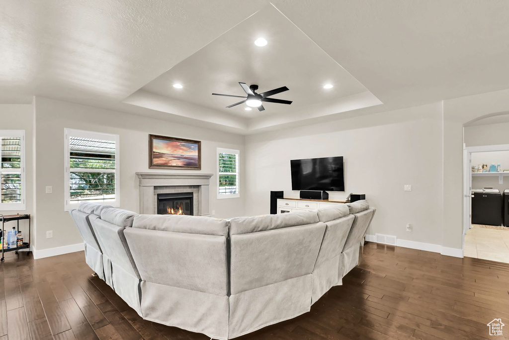 Living area with ceiling fan, a glass covered fireplace, dark wood finished floors, baseboards, and a tray ceiling