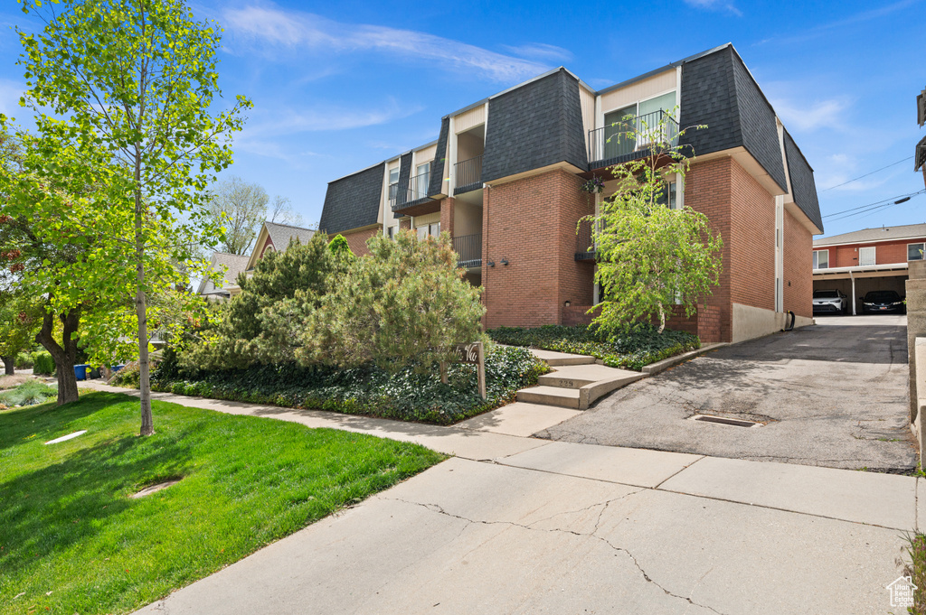 View of property exterior featuring a lawn, brick siding, mansard roof, a balcony, and a shingled roof