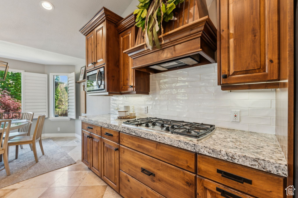 Kitchen with brown cabinets, appliances with stainless steel finishes, baseboards, custom range hood, and backsplash
