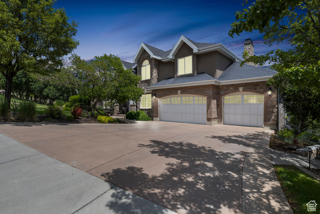 View of front of house featuring a shingled roof, an attached garage, stone siding, a chimney, and concrete driveway
