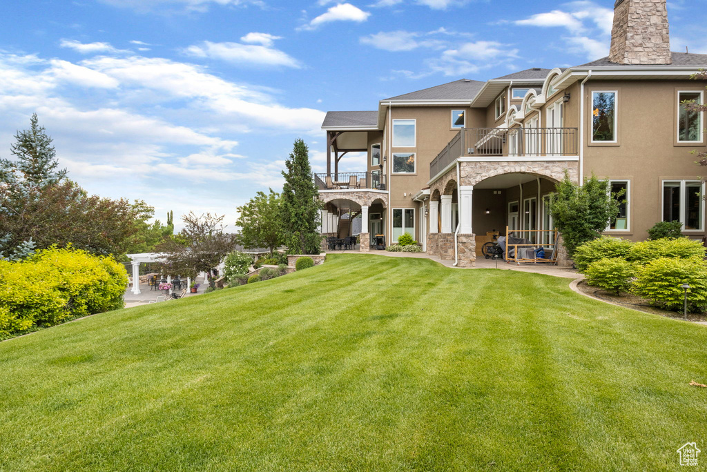 Rear view of house featuring a balcony, stone siding, a patio, stucco siding, and a lawn
