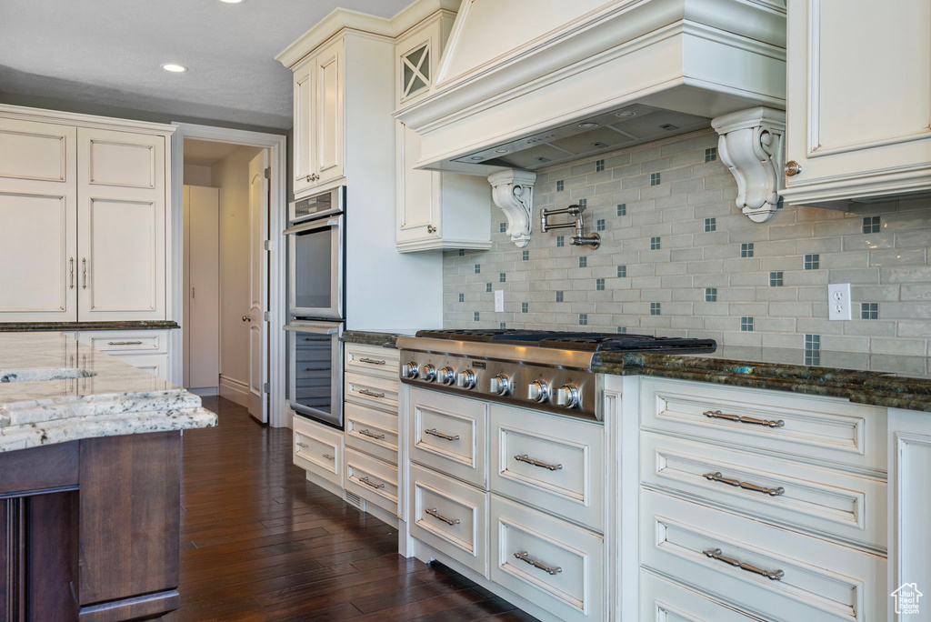 Kitchen featuring custom exhaust hood, dark wood-style floors, dark stone countertops, backsplash, and stainless steel appliances