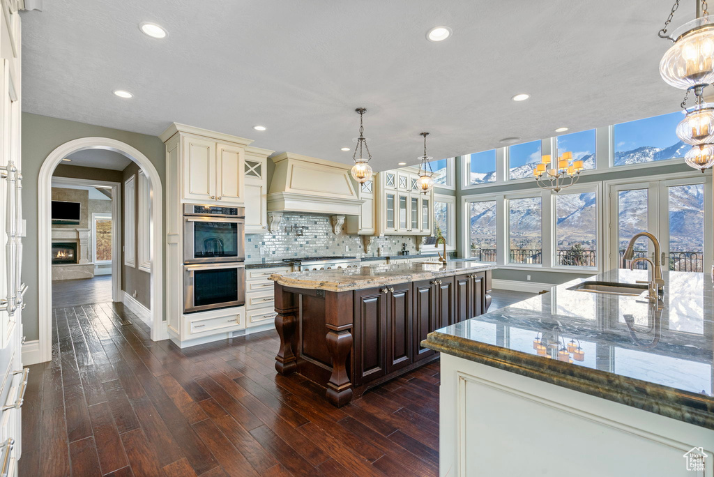 Kitchen featuring a kitchen island with sink, baseboards, stainless steel double oven, custom range hood, and a sink