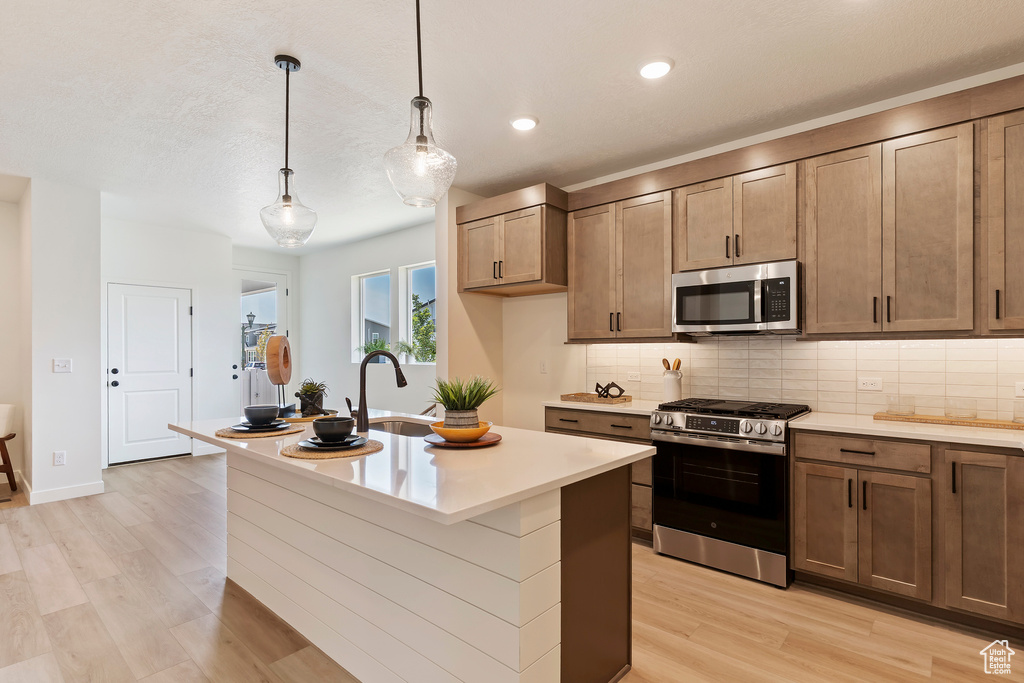 Kitchen featuring appliances with stainless steel finishes, light wood-style floors, tasteful backsplash, light countertops, and an island with sink
