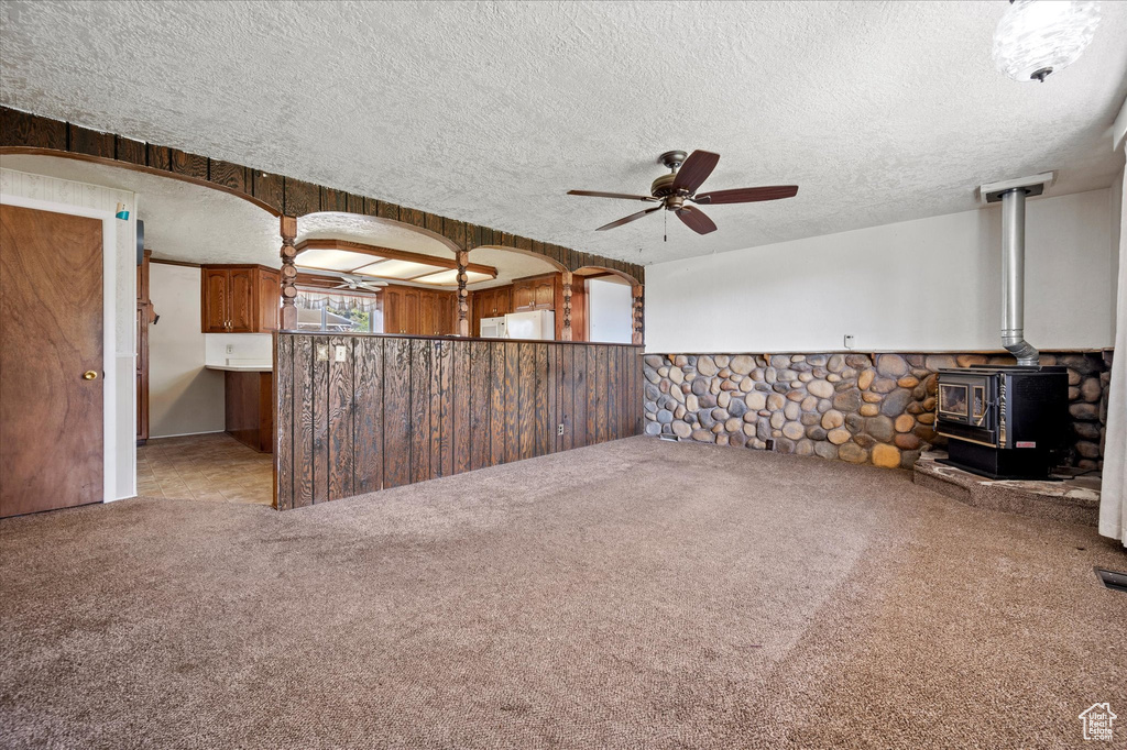 Unfurnished living room with a wood stove, carpet floors, a textured ceiling, a wainscoted wall, and a ceiling fan