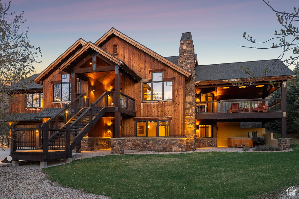 Back of property at dusk featuring a balcony, stone siding, a chimney, stairs, and a yard