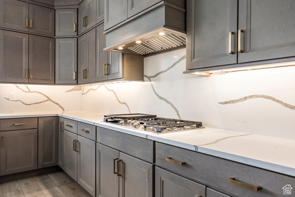 Kitchen featuring gray cabinets, tasteful backsplash, stainless steel gas cooktop, custom exhaust hood, and light wood finished floors