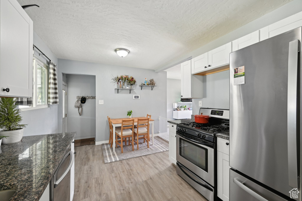 Kitchen with a textured ceiling, appliances with stainless steel finishes, light wood-style floors, dark stone counters, and white cabinetry