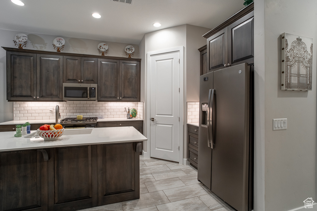 Kitchen with dark brown cabinetry, appliances with stainless steel finishes, a sink, light countertops, and backsplash