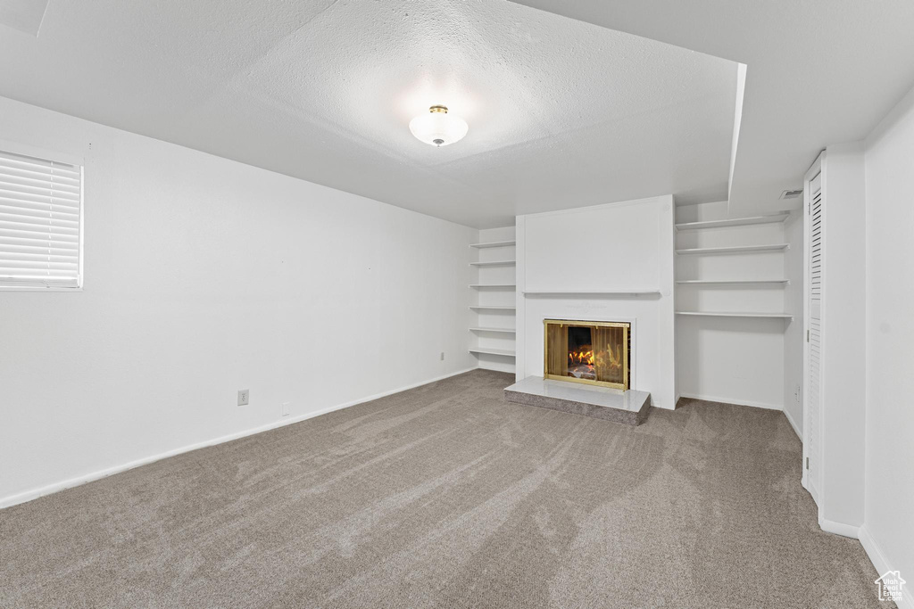 Unfurnished living room featuring a textured ceiling, built in shelves, baseboards, a warm lit fireplace, and carpet flooring
