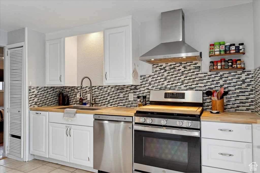 Kitchen featuring butcher block counters, wall chimney exhaust hood, white cabinets, a sink, and appliances with stainless steel finishes