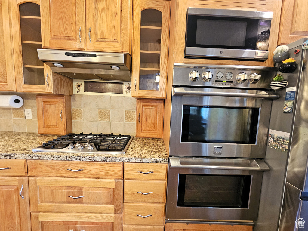 Kitchen featuring under cabinet range hood, stainless steel appliances, glass insert cabinets, and tasteful backsplash