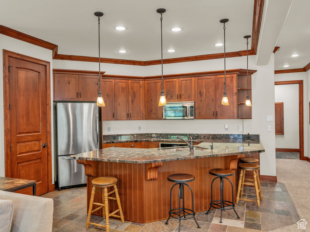 Kitchen featuring hanging light fixtures, appliances with stainless steel finishes, a breakfast bar area, dark stone counters, and ornamental molding