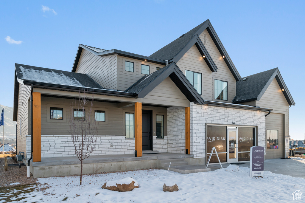 Snow covered back of property with covered porch, roof with shingles, and stone siding