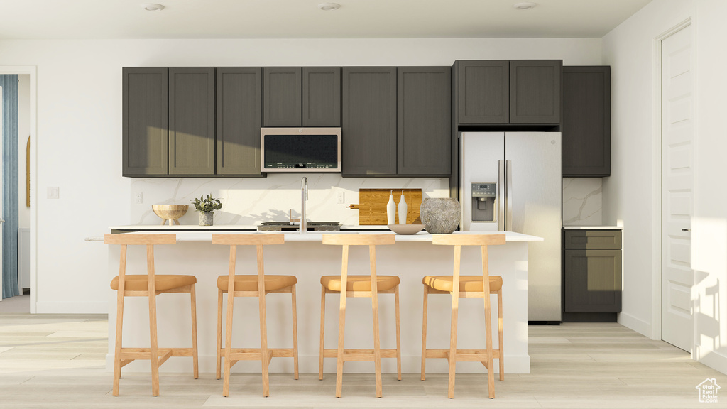 Kitchen featuring a kitchen island with sink, light wood-type flooring, light countertops, stainless steel microwave, and refrigerator with ice dispenser