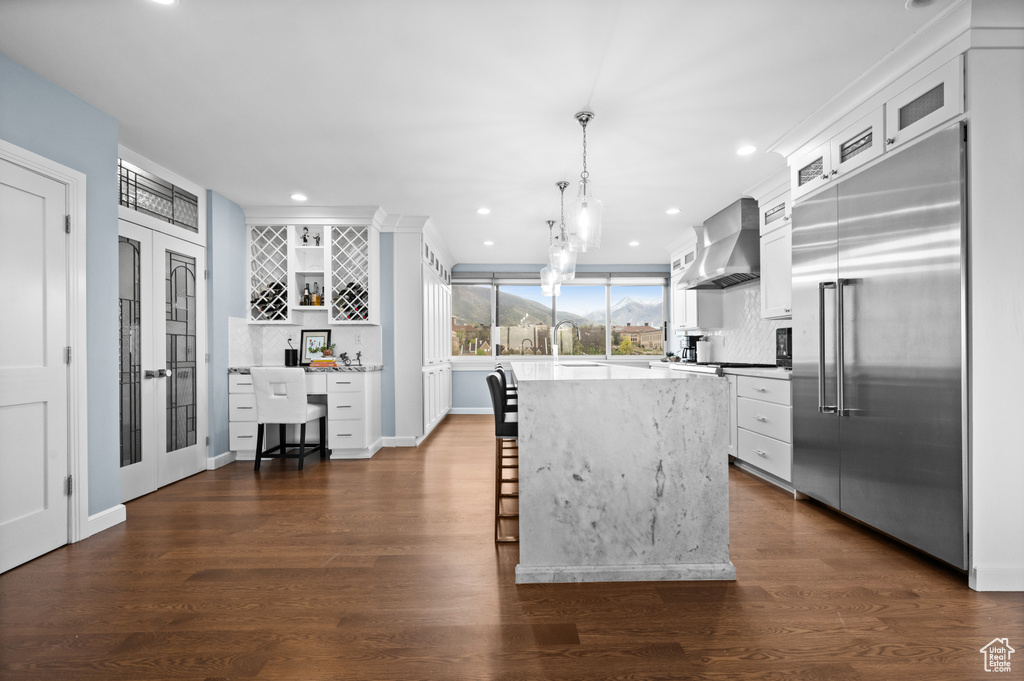 Kitchen featuring a kitchen bar, built in refrigerator, wall chimney exhaust hood, white cabinets, and tasteful backsplash