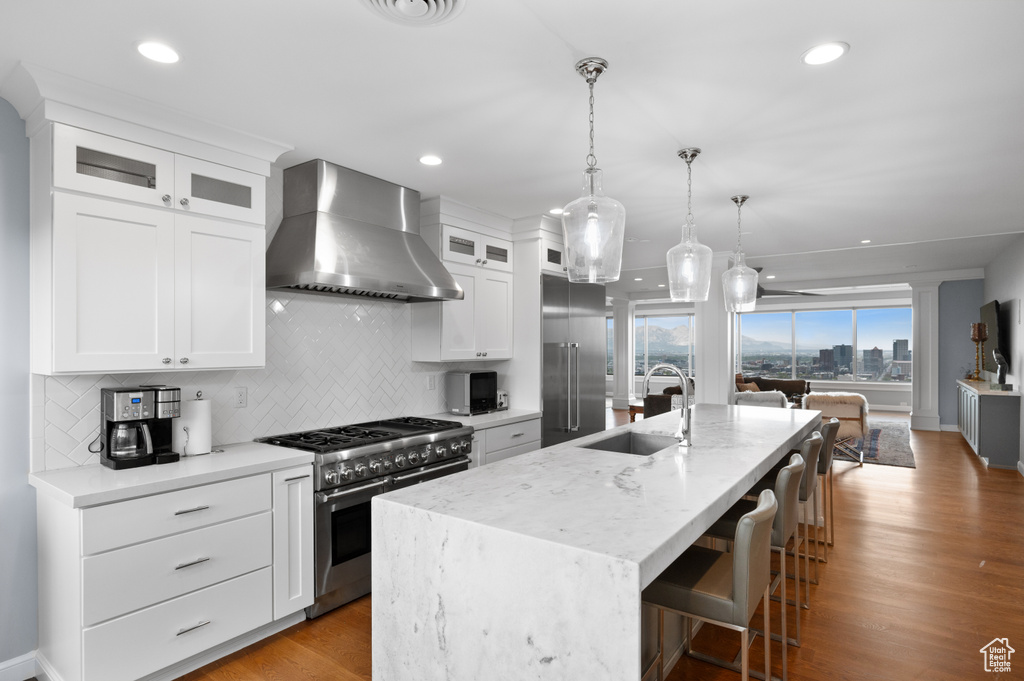 Kitchen featuring a sink, a kitchen island with sink, wall chimney exhaust hood, backsplash, and premium appliances