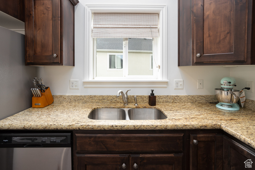 Kitchen featuring stainless steel dishwasher, a sink, dark brown cabinets, and light stone countertops
