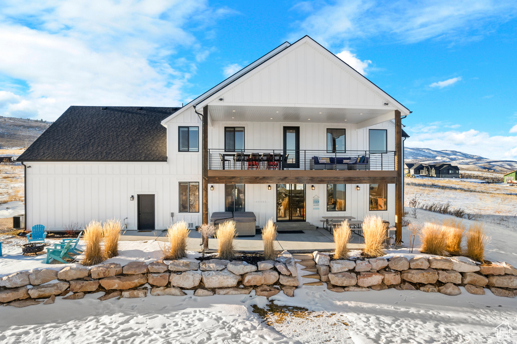 Snow covered back of property with roof with shingles, board and batten siding, a mountain view, a patio area, and a balcony