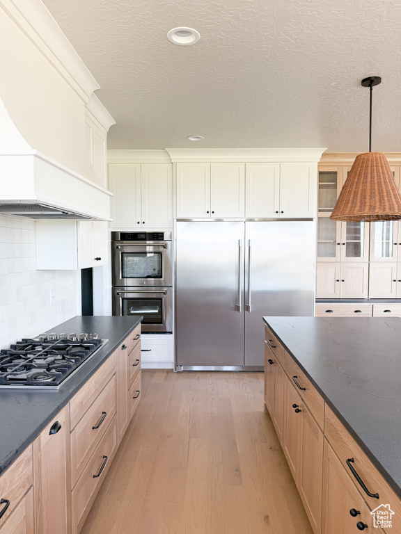 Kitchen with decorative backsplash, decorative light fixtures, appliances with stainless steel finishes, light wood finished floors, and custom range hood