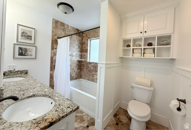 Full bathroom with toilet, shower / tub combo with curtain, double vanity, and a wainscoted wall