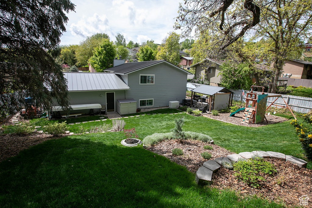 Back of property featuring an outdoor fire pit, a fenced backyard, a playground, a yard, and metal roof