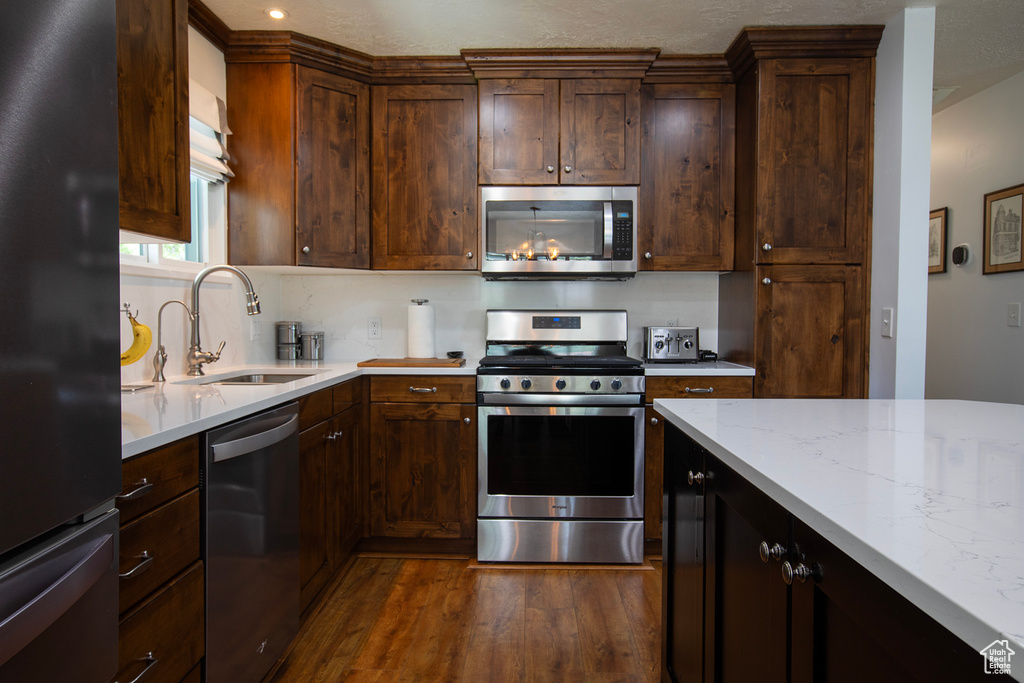 Kitchen with dark brown cabinetry, a sink, light stone counters, dark wood-style floors, and stainless steel appliances