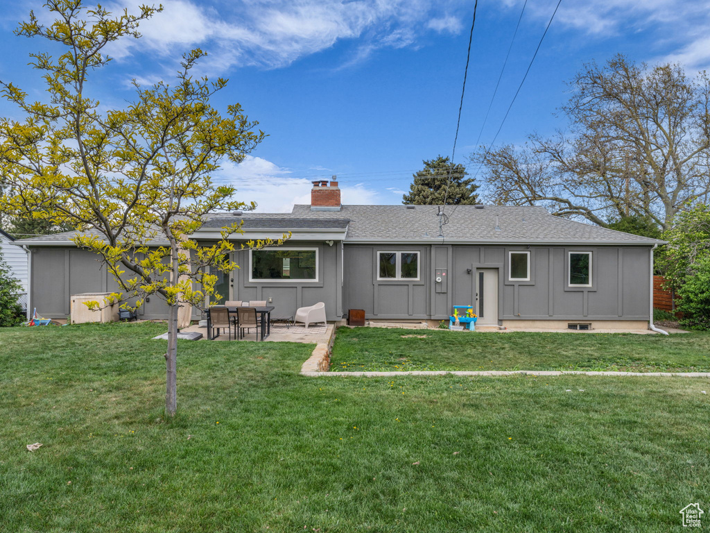 Rear view of house featuring board and batten siding, a chimney, roof with shingles, a lawn, and a patio