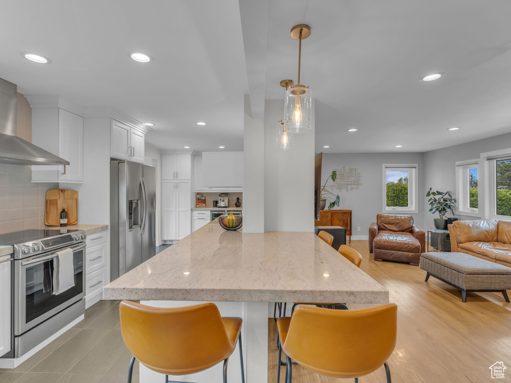 Kitchen featuring open floor plan, wall chimney range hood, recessed lighting, white cabinetry, and stainless steel appliances