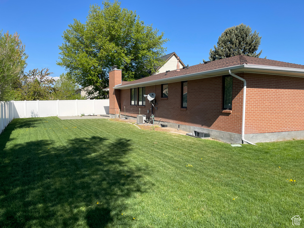 Back of property featuring central AC, a fenced backyard, a chimney, brick siding, and a yard