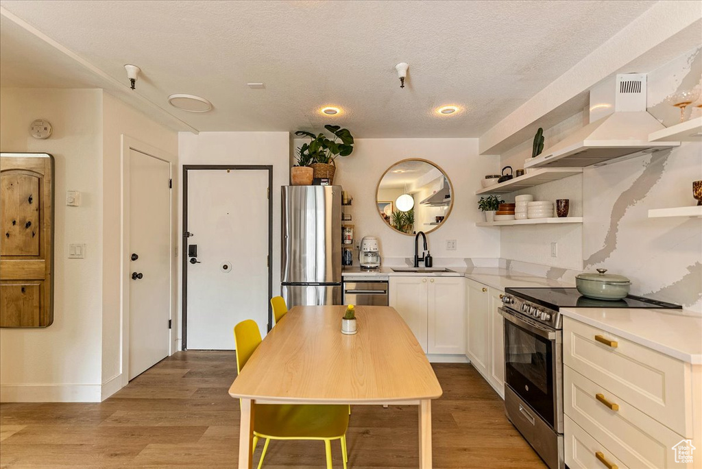 Kitchen featuring wall chimney range hood, open shelves, a sink, light wood-style floors, and stainless steel appliances