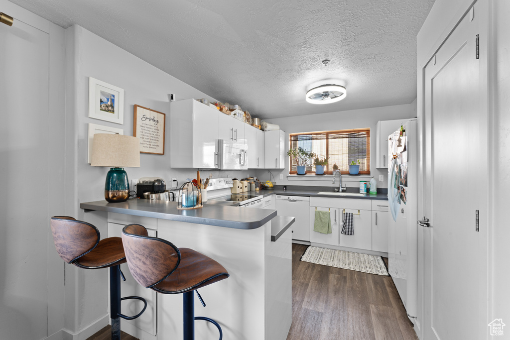 Kitchen featuring white cabinets, a sink, white appliances, dark wood finished floors, and a peninsula