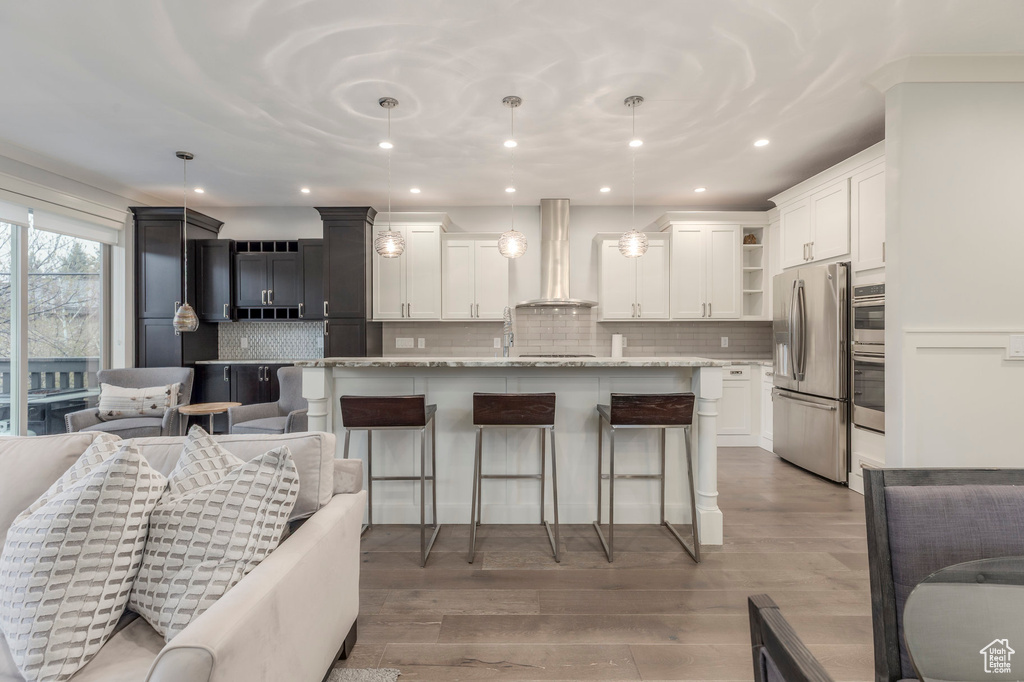 Kitchen featuring wall chimney range hood, open shelves, an island with sink, stainless steel appliances, and tasteful backsplash