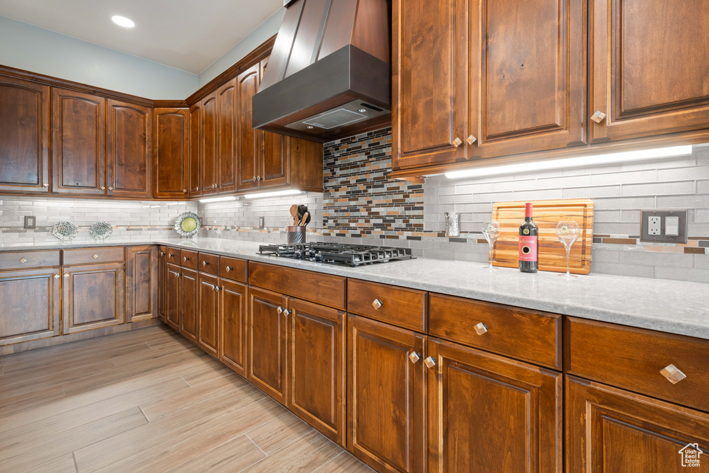 Kitchen with light stone countertops, tasteful backsplash, wall chimney exhaust hood, stainless steel gas stovetop, and light wood-type flooring
