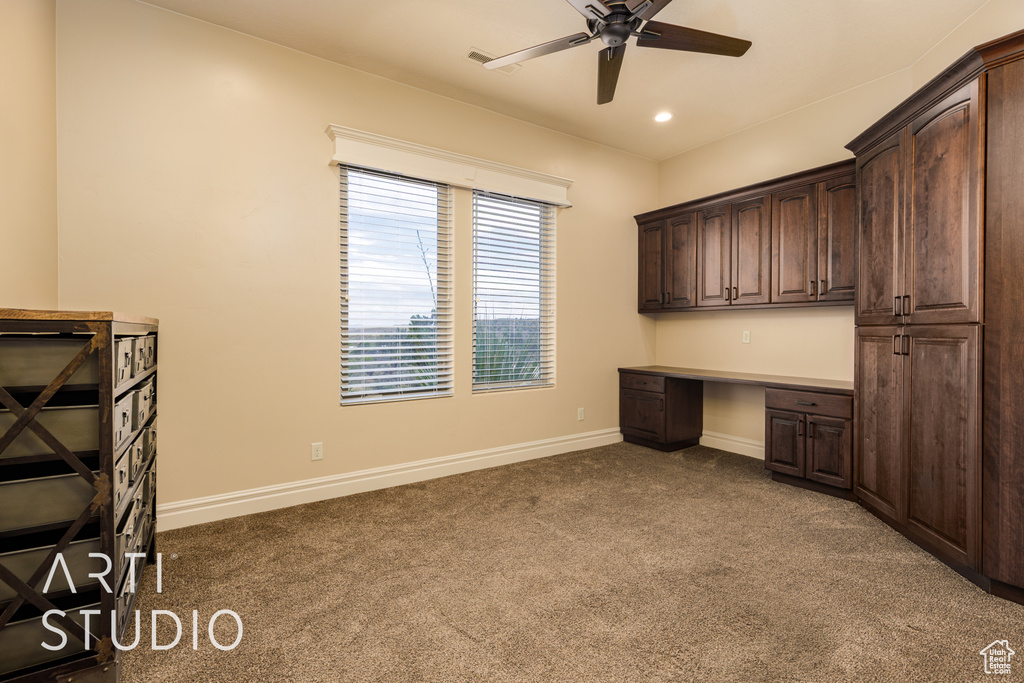 Unfurnished office with dark colored carpet, a ceiling fan, baseboards, and built in desk