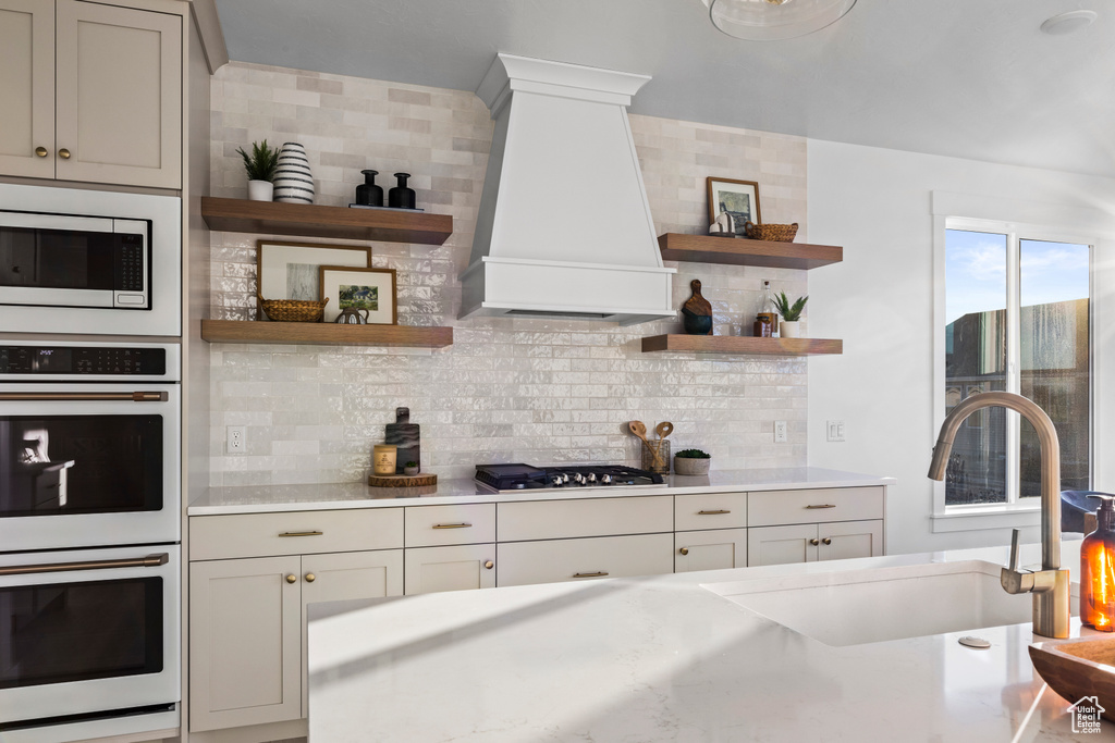 Kitchen with open shelves, double oven, stainless steel microwave, custom range hood, and tasteful backsplash