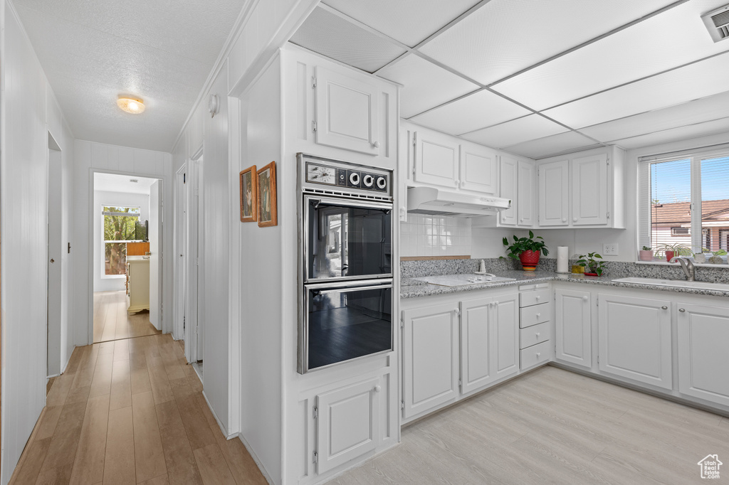 Kitchen featuring double oven, white cabinetry, under cabinet range hood, a sink, and light wood finished floors