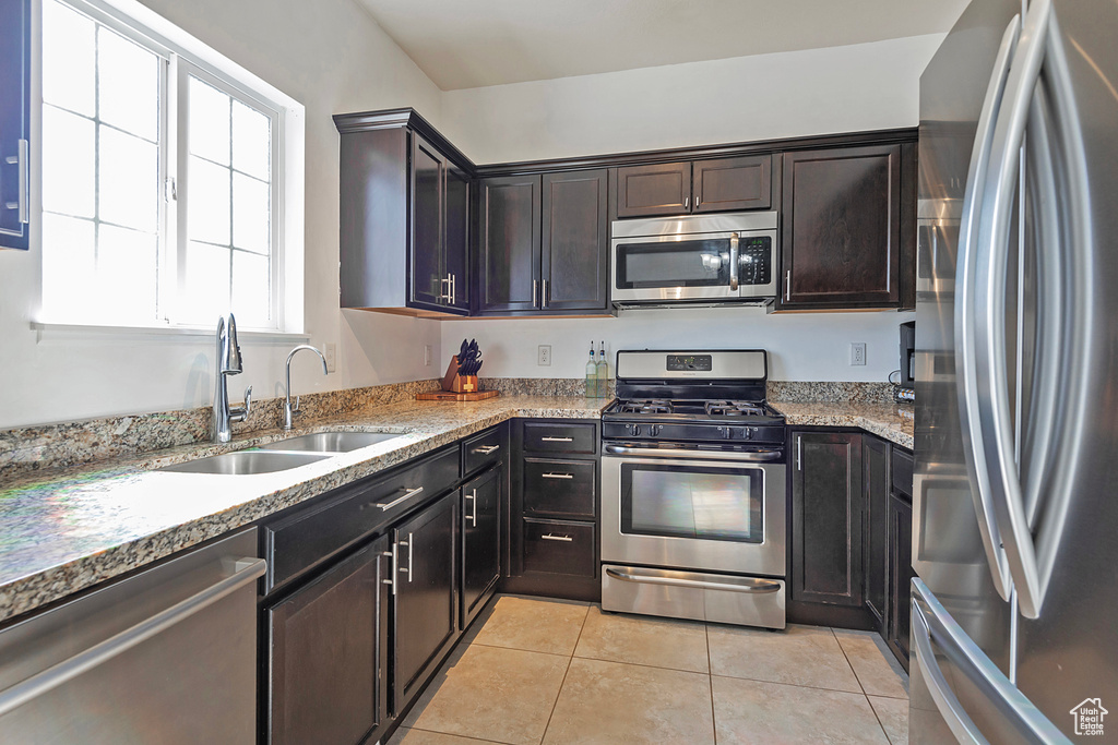 Kitchen with light tile patterned flooring, dark brown cabinetry, appliances with stainless steel finishes, light stone counters, and a sink
