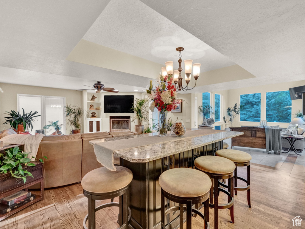 Dining room featuring a fireplace, a raised ceiling, a textured ceiling, light wood finished floors, and ceiling fan with notable chandelier