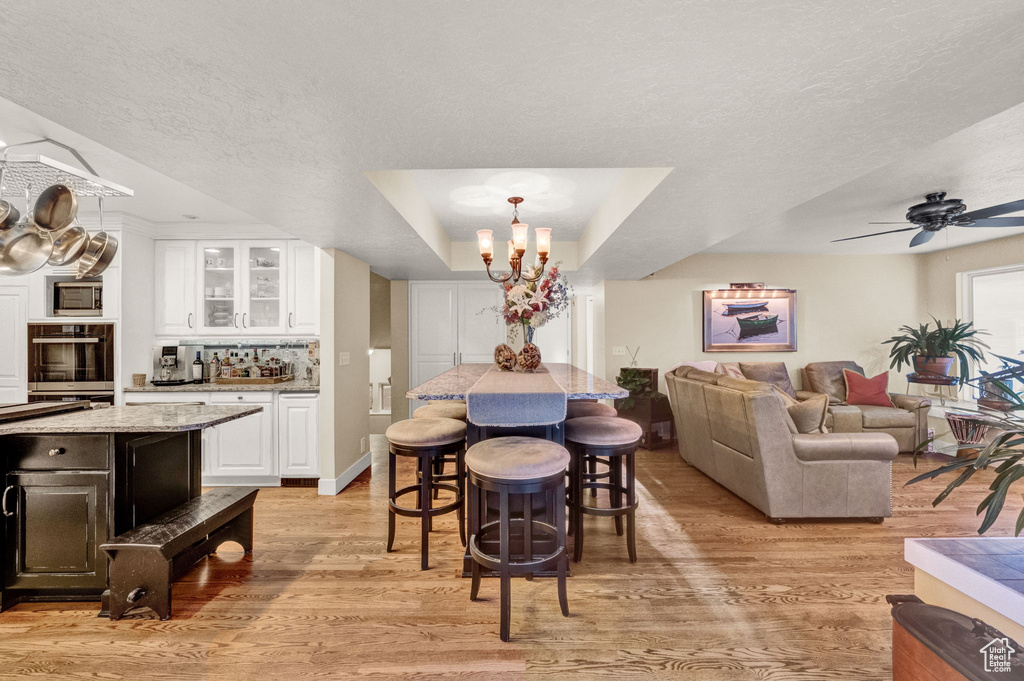 Dining area with a raised ceiling, ceiling fan with notable chandelier, light wood finished floors, and a textured ceiling