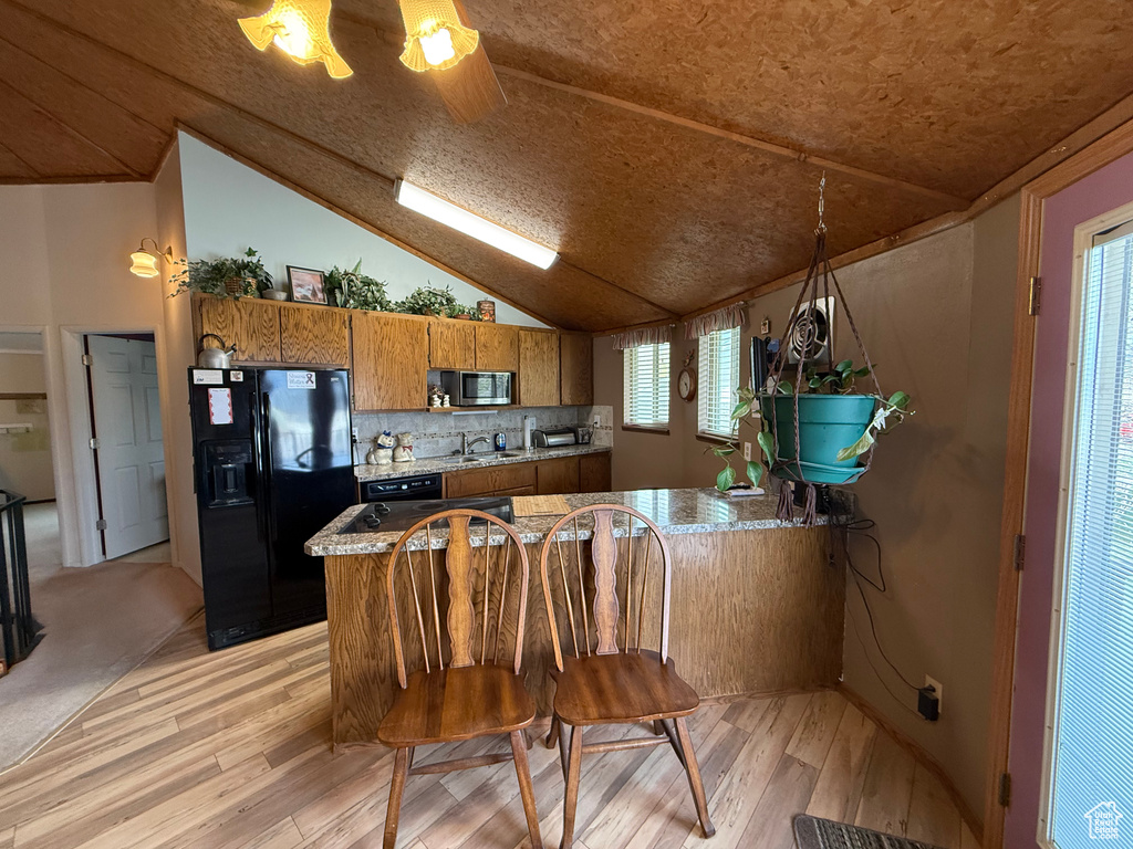 Kitchen featuring vaulted ceiling, light wood-type flooring, black fridge with ice dispenser, brown cabinets, and stainless steel microwave