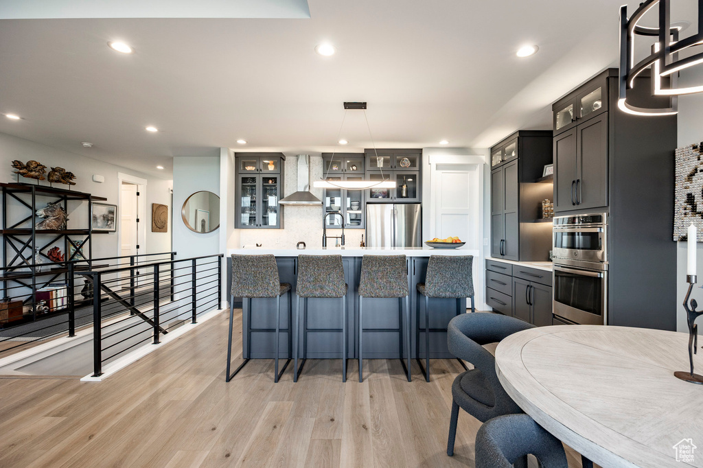 Kitchen featuring a breakfast bar area, wall chimney range hood, light countertops, light wood-style flooring, and stainless steel appliances