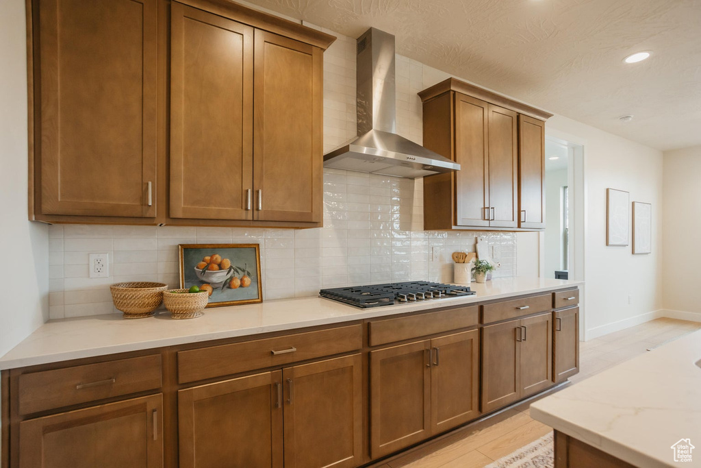Kitchen with wall chimney range hood, stainless steel gas cooktop, backsplash, and light wood-style flooring