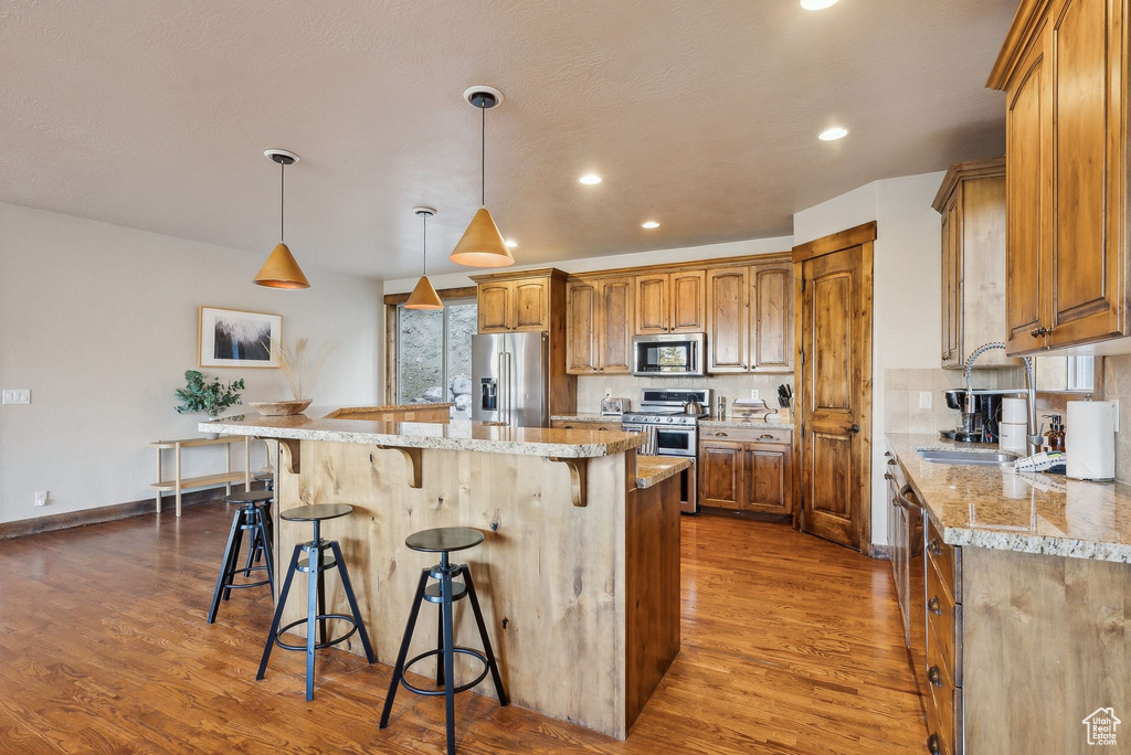 Kitchen featuring appliances with stainless steel finishes, a kitchen breakfast bar, brown cabinetry, a kitchen island, and dark wood-style floors