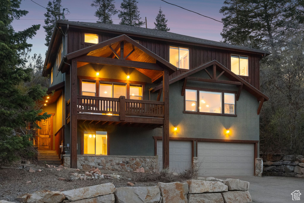 Back of house at dusk featuring a garage, a balcony, stone siding, stucco siding, and concrete driveway