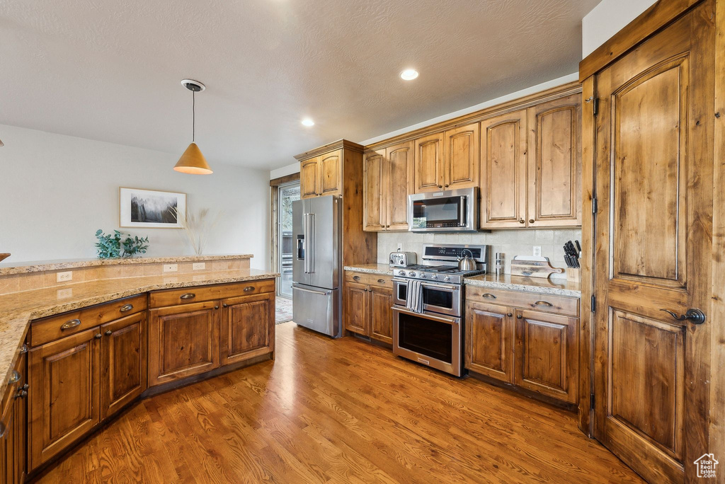 Kitchen with pendant lighting, appliances with stainless steel finishes, light stone countertops, brown cabinets, and wood finished floors